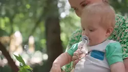 دانلود فوتیج نوزاد با پستانک در حال لمس برگ درخت در پارک