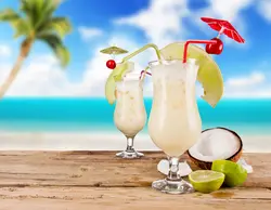 دانلود تصویر شاتراستوک دو لیوان اسموتی با چتر تزئینی و نارگیل و لیمو روی میز چوبی در ساحل کد۱۲۹۲۳۲۱۵۲۴