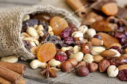 عکس با کیفیت کیسه کوچک کنفی حاوی انواع مغزهای آجیل شامل پسته ، فندق ، بادام و بادام هندی به همراه کشته و کشمش ریخته شده بر روی سطح میز چوبی و چوب های دارچین در کنار آن