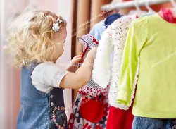 عکس با کیفیت دختر کوچک یا دختر بچه با موهای بلوند در حال بررسی لباس های بچه گانه داخل رگال در بوتیک