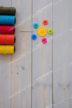 عکس با کیفیت قرقره های نخ های رنگی در کنار دکمه های رنگی و سوزن های چیده شده روی میز چوبی