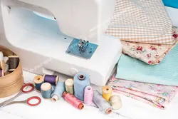 عکس با کیفیت ابزار خیاطی و جعبه نخ های رنگارنگ و پارچه های گل دار و چهارخانه در کنار چرخ خیاطی سفید رنگ