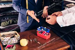 عکس با کیفیت خیاط آقایان در حال سفارش گیری از مشتری در کنار میز و ابزار خیاطی و طاقه های پارچه