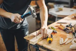 عکس با کیفیت نجار در حال کوبیدن میخ با چکش بر روی چوب در کنار ابزار کاتر ، نقاله ، رنده ، کولیس و گیره ویژه استفاده در امور تبلیغاتی و تجاری طراحی کاتالوگ ، بروشور و تراکت با موضوع نجاری ، صنایع چوب و ساخت میز ، مبل و صندلی